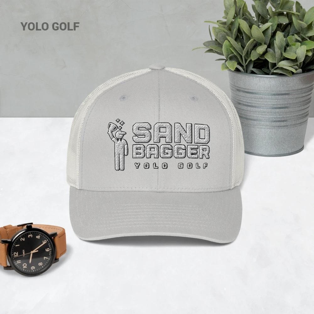 Light gray Sandbagger trucker hat displayed on a table with a watch and plant, highlighting its 