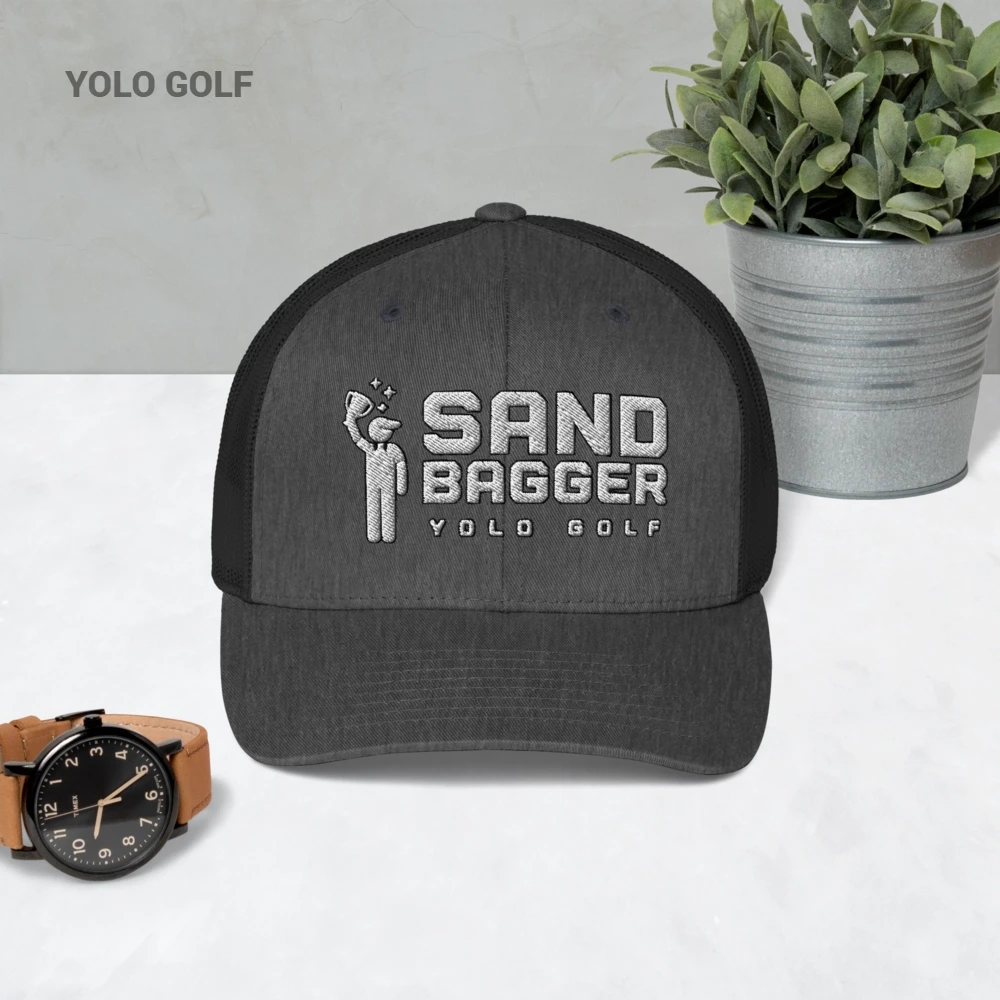 Charcoal gray Sandbagger trucker hat displayed on a table with a watch and plant, showcasing its 